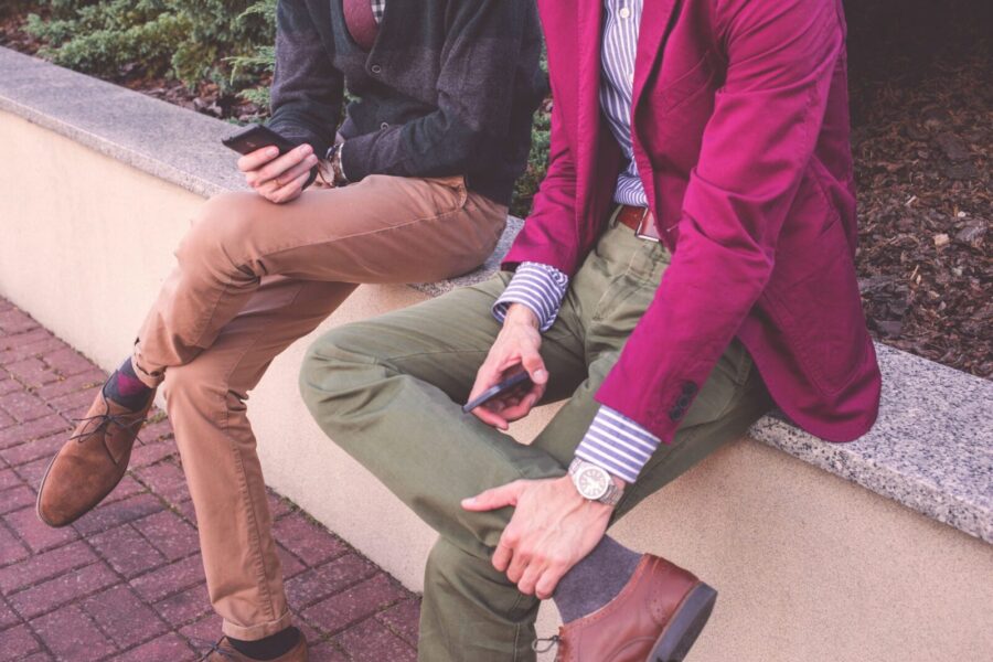 Two men casually dressed in fashion jackets, sitting outdoors and using smartphones, showcasing friendship and modern lifestyle.